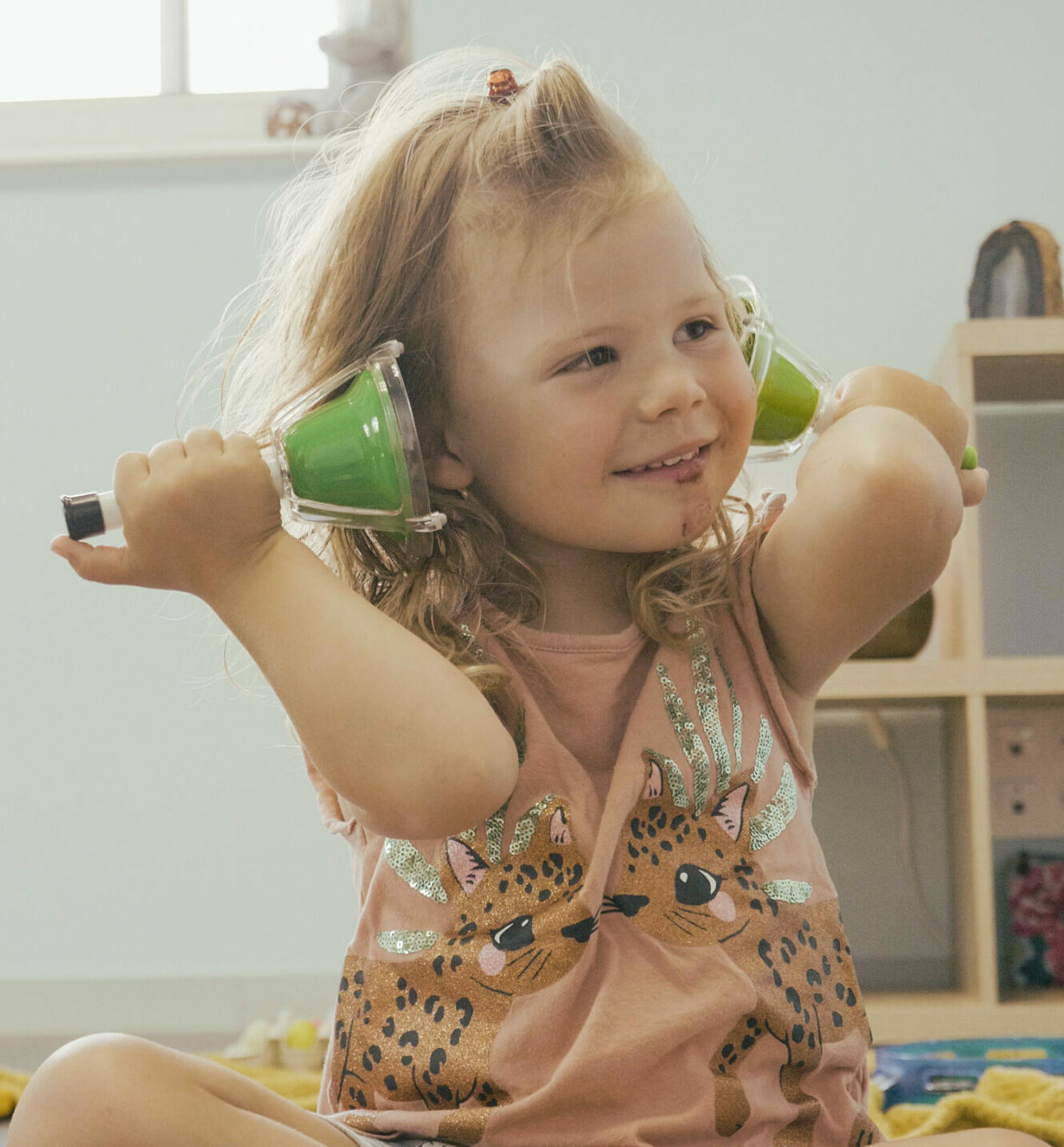 Photo d'un enfant ris 3/4 profil qui tient des cloches à pouce dans chaque main de part et d'autres de ses oreilles. Il a un grand sourire et semble regarder sur sa gauche quelque chose avec attention.